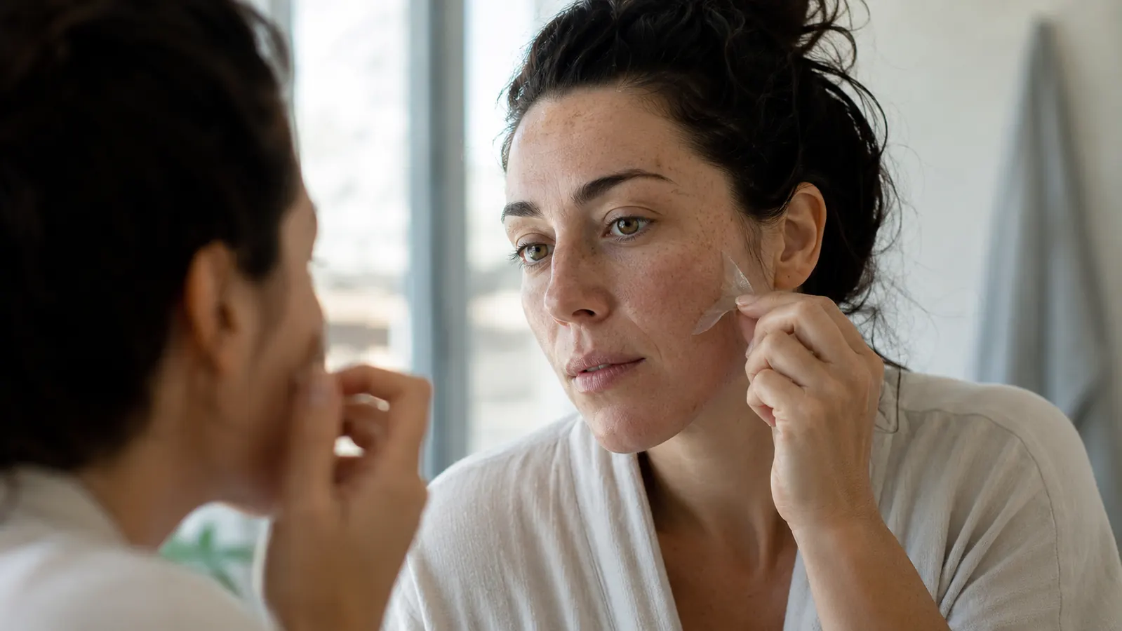 Person in the morning peeling a small patch off their cheek in natural daylight before getting ready