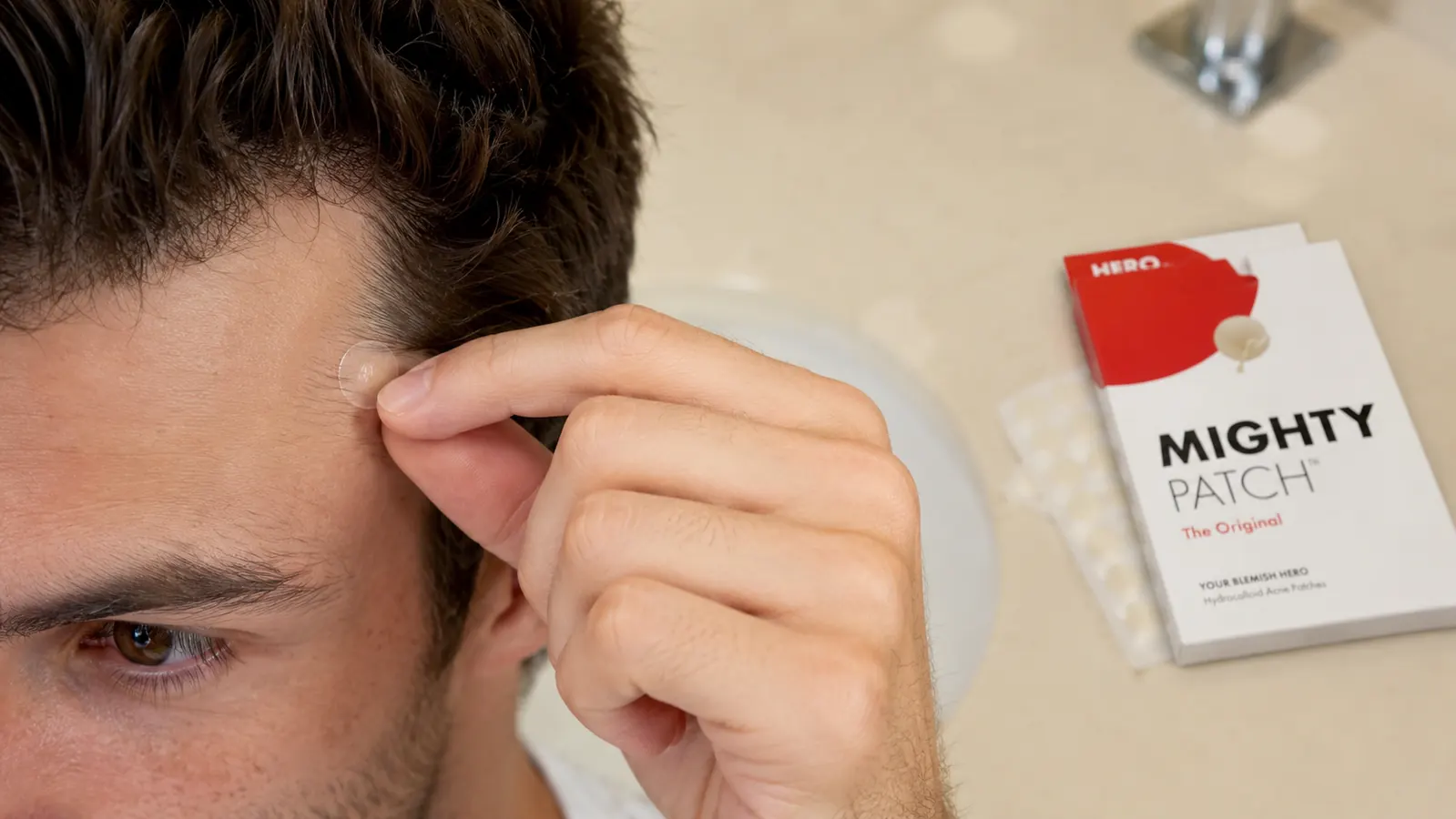 Hand pressing a small round hydrocolloid patch onto a breakout near the hairline and temple