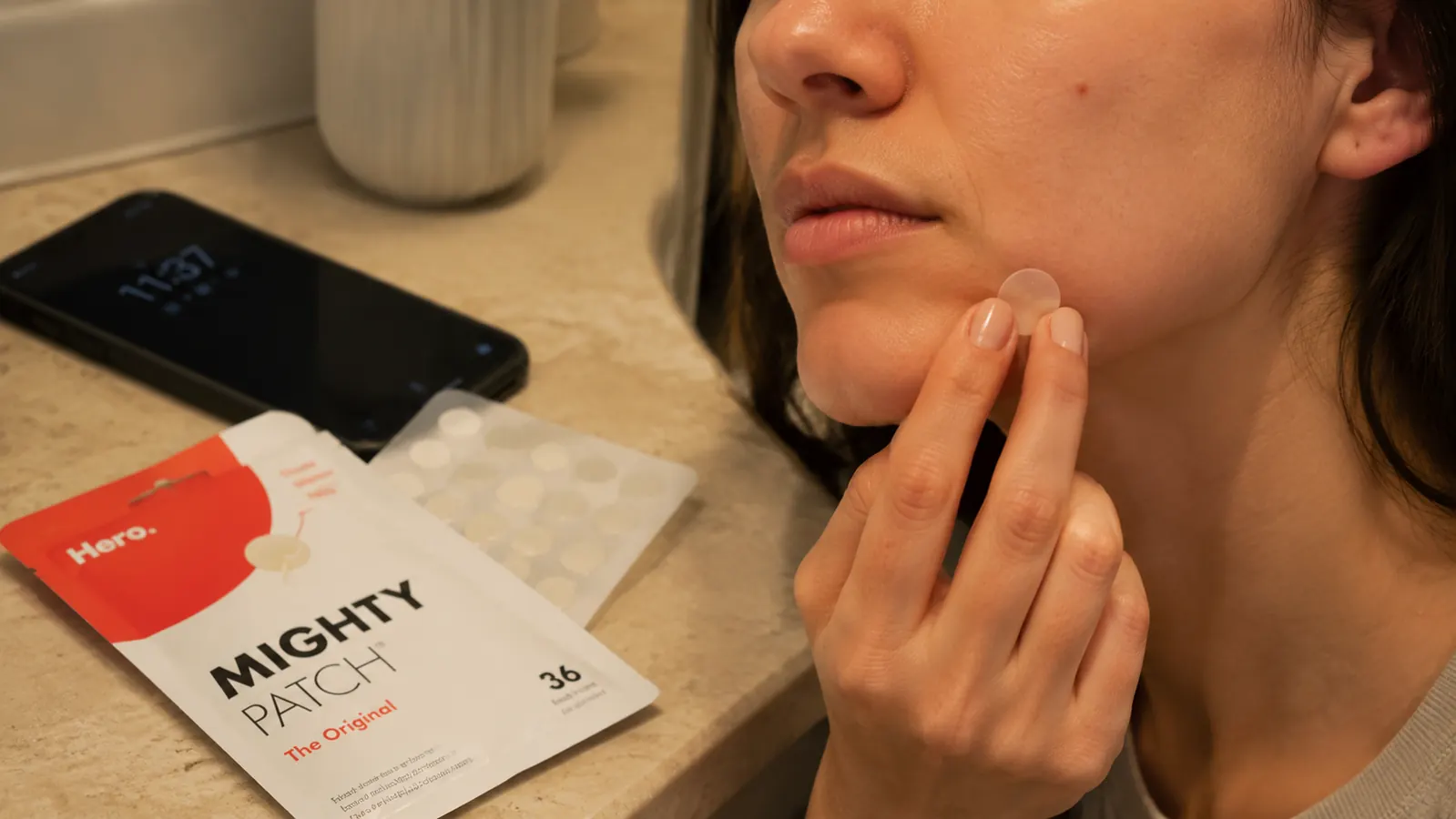 Hand pressing a small clear hydrocolloid patch onto a red bump near the jawline in a bathroom